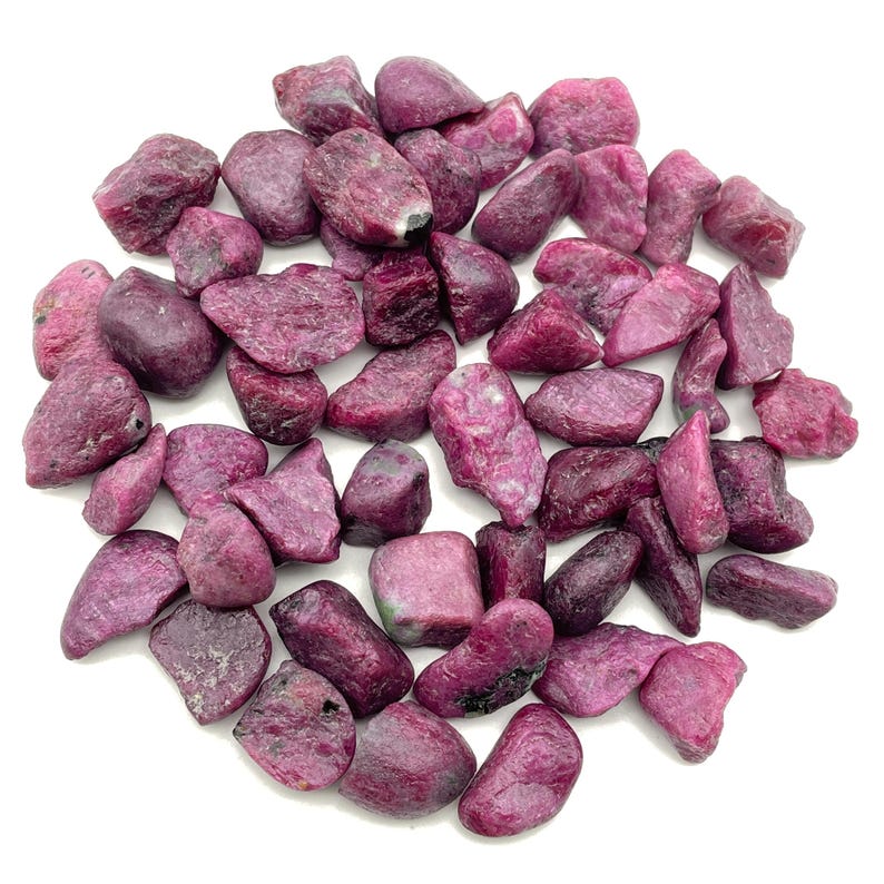 Peut inclure: Un ensemble de pierres pr&eacute;cieuses de rubis brutes de formes et de tailles vari&eacute;es. Les pierres pr&eacute;sentent une gamme de teintes rouge fonc&eacute; et rose, avec une surface rugueuse et textur&eacute;e. Les pierres sont dispers&eacute;es sur un fond blanc.