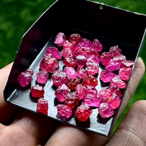 May include: A collection of small, raw, vibrant pinkish-red gemstones scattered across a black metal scoop. The stones exhibit irregular, faceted, and crystalline shapes with varying degrees of translucency and deep magenta to bright crimson hues.