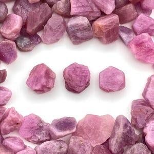 Könnte beinhalten: Eine Ansammlung roher, ungeschliffener Rubin-Edelsteine in verschiedenen Rosa- und Lilatönen. Die Edelsteine sind auf einer weißen Oberfläche verstreut, einige in einer Reihe angeordnet. Die raue Textur und die natürlichen Formen der Steine sind sichtbar.