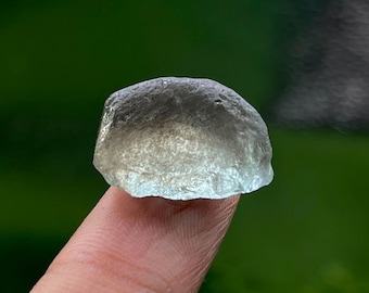 May include: A translucent, light blue-green gemstone with a rough, textured surface. The stone is held up by a finger, with a blurred green background. The gemstone appears to be a natural, uncut specimen.