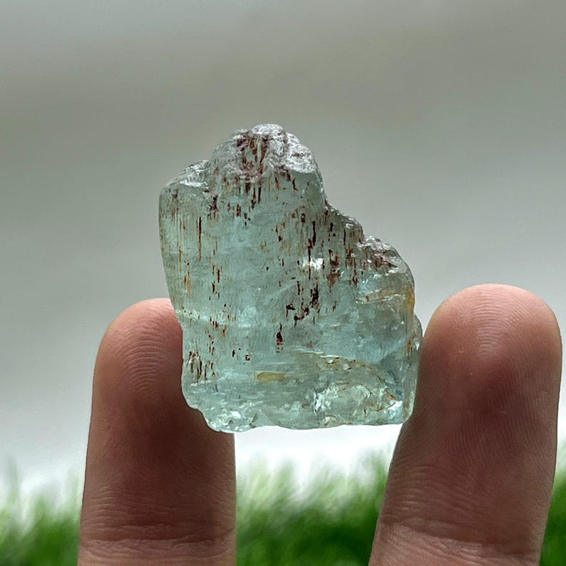 May include: A rough-cut aquamarine gemstone with a pale blue hue and brown inclusions. The natural crystal is held between two fingers, showcasing its irregular shape and translucent quality. The gemstone is a mineral specimen.