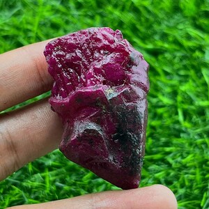May include: A rough, uncut ruby gemstone with a deep red hue and a textured surface. The natural stone displays dark patches and irregular edges, showcasing its raw, unpolished state. The ruby is held against a backdrop of green grass.