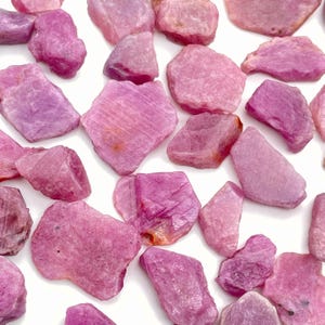 May include: A selection of raw ruby gemstones in shades of pink and magenta. The uncut stones have irregular shapes and textures, scattered on a white surface. The gemstones vary in size and colour intensity.