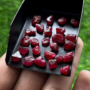 Pode incluir: Uma coleção de pequenas pedras preciosas de rubi brutas e vermelho-escuras espalhadas por uma pá de metal preta. Cada pedra apresenta superfícies irregulares e facetadas com níveis variados de translucidez e uma cor carmesim vibrante e saturada.