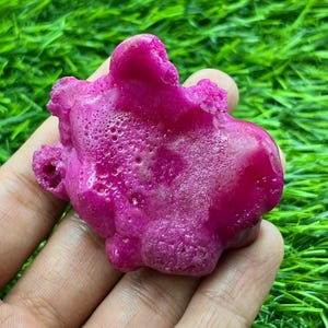 May include: A fuchsia-coloured gemstone with an irregular shape and textured surface. The stone is held in a hand against a backdrop of green grass, highlighting its natural beauty and unique form.