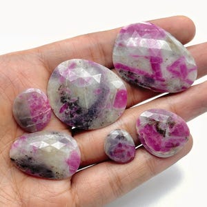 May include: A selection of faceted ruby gemstones in various oval and rounded shapes. The gemstones display a mix of pink, white, and grey hues, with visible internal patterns. The stones are held in a hand.
