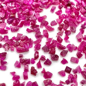 Puede incluir: Una dispersión de piedras preciosas de rubí en bruto, sin tallar, de diversas formas y tamaños. Las piedras son de un color rojo-rosado intenso, con un aspecto ligeramente translúcido. La textura rugosa de las piedras es visible.
