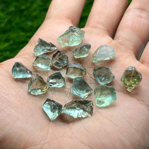 Puede incluir: Una colección de pequeñas piedras preciosas en bruto, translúcidas, de color verde menta y amarillo pálido, con formas irregulares y dentadas y superficies facetadas. Estas piedras minerales sueltas muestran diversos grados de claridad.