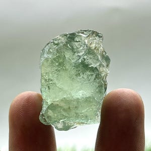 May include: A translucent, light green raw fluorite crystal. The rough-hewn stone has a textured surface and is held between two fingers. The crystal's color varies from pale to medium green, with some areas appearing almost clear.