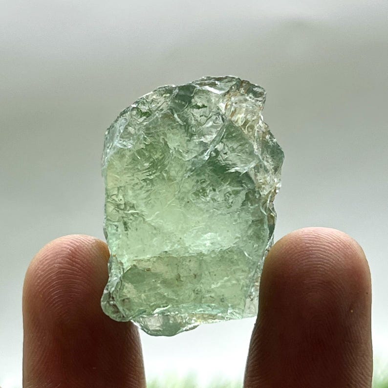 May include: A translucent, light green raw fluorite crystal. The rough-hewn stone has a textured surface and is held between two fingers. The crystal's color varies from pale to medium green, with some areas appearing almost clear.
