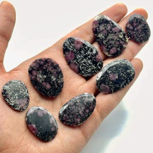 Puede incluir: Una colección de piedras preciosas ovaladas con un patrón moteado gris oscuro y negro, intercaladas con inclusiones rojas y rosas vibrantes. Las piedras se sostienen en una mano sobre un fondo blanco, mostrando sus superficies pulidas.