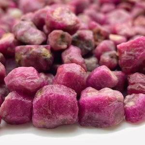 Puede incluir: Primer plano de un montón de piedras preciosas de rubí en bruto sin cortar. Las piedras son de un color rojo rosado intenso, con una superficie ligeramente irregular y texturizada. Las piedras preciosas están agrupadas, creando una disposición densa y natural.