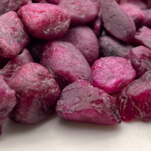 Puede incluir: Primer plano de un montón de piedras preciosas de rubí en bruto. Las piedras varían en forma y tamaño, con un tono rojo-púrpura intenso. Se aprecia la textura rugosa y las formas naturales de las piedras, lo que sugiere que no están pulidas.