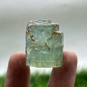 May include: A raw, light blue aquamarine crystal with a rectangular shape. The gemstone has a translucent quality with visible internal structures and some brown inclusions. The crystal is held between two fingers, showcasing its natural form and color.