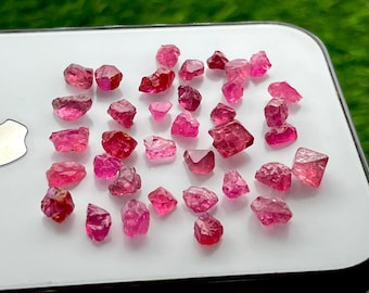 May include: A collection of small, raw, faceted-style pinkish-red gemstones scattered across a smooth white surface. These translucent stones exhibit irregular, angular shapes with varying depths of red and pink hues throughout their crystalline structures.