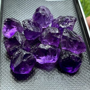 Puede incluir: Un conjunto de diez piedras de amatista en bruto de color púrpura intenso con superficies irregulares y facetadas. Los cristales translúcidos y dentados están agrupados sobre una superficie negra texturizada con el texto impreso PG 13/14 2 RH.