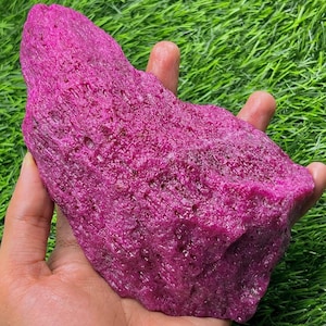 Könnte beinhalten: Ein großer, ungeschliffener Rubin-Edelstein in tiefem Magenta. Das rohe Mineral wird in einer Hand gehalten und zeigt seine unregelmäßige Form und strukturierte Oberfläche. Der Rubin ist ein natürliches, ungeschnittenes Exemplar.