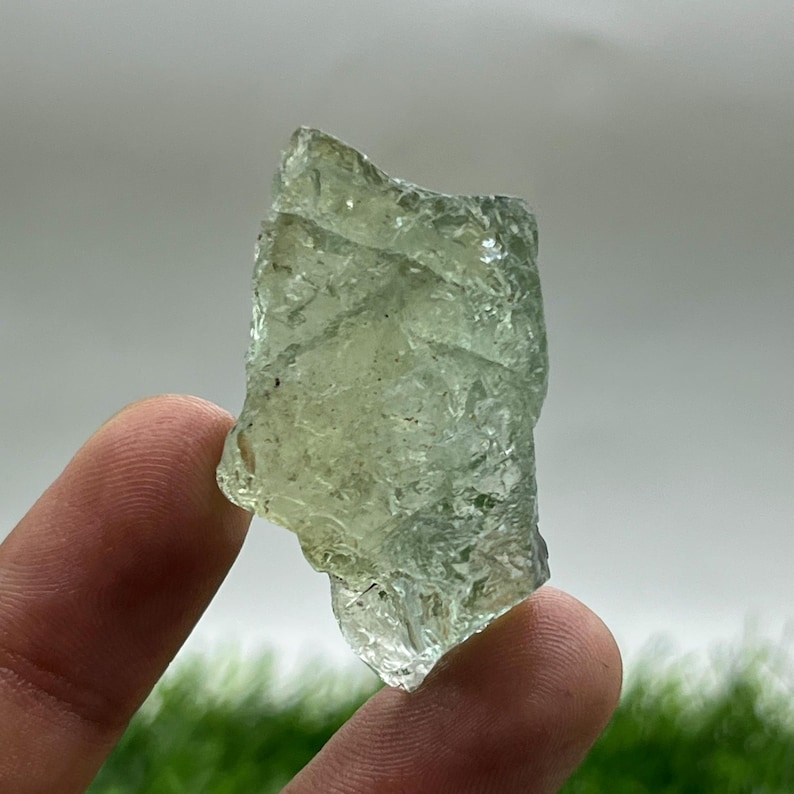 May include: A translucent, pale green rough gemstone with a crystalline structure. The irregular shape of the stone is held between two fingers, with a blurred green and white background.
