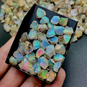 May include: A collection of raw opals in a black tray. The opals display a variety of colors, including blue, green, and pink, with a rough, natural texture. The tray is held in a hand, with more opals visible in the background.