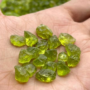 May include: A selection of raw peridot gemstones, displaying a vibrant, translucent green hue. The uncut stones have a rough, natural texture and are presented in a hand, highlighting their natural beauty. The peridot stones are of various sizes.