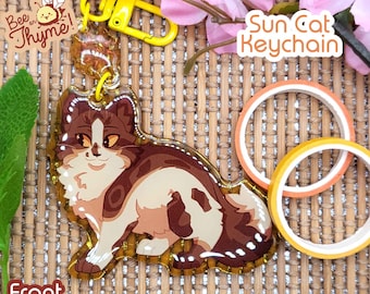 May include: A brown, white, and orange cat keychain with a yellow glitter background. The keychain is shaped like a cat sitting down. The text "Sun Cat Keychain" is printed on a white background above the cat.