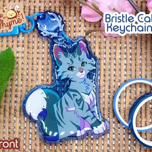 以下が含まれることがあります： キラキラ光る青いグレーの猫のキーホルダー。キーホルダーは、尻尾を脚に巻き付けた座っている猫の形をしています。猫は白い胸と額に白い斑点があります。キーホルダーは青と銀の金属製のリングに取り付けられています。