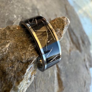 May include: A black and silver ring with a unique design. The ring features a dark band with white streaks and a silver border. The ring is displayed on a textured, gray stone, showcasing its details and craftsmanship.