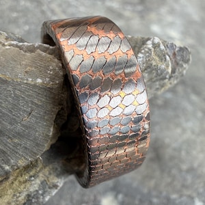May include: A wide, textured metal bracelet with a pattern of overlapping scales. The bracelet features a combination of copper and silver tones, creating a striking contrast. The bracelet is displayed on a gray stone surface.