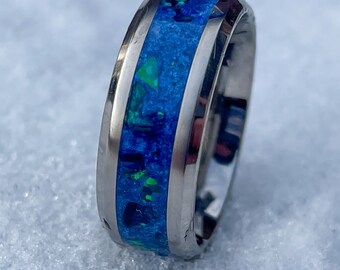 May include: A silver-colored ring with a central band of blue and green opal. The ring has a polished finish and a smooth, rounded design. The opal has a vibrant, iridescent quality.