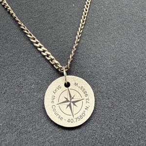 May include: Silver-coloured necklace with a circular pendant. The pendant has a compass rose design and the words "Stay the Course" and coordinates. The chain is a curb style.