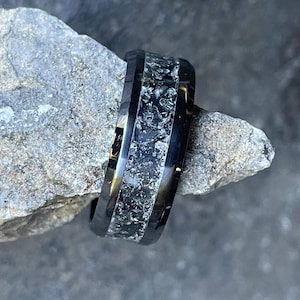 Può includere: Un anello nero e argento con una fascia testurizzata. La fascia è realizzata in metallo scuro con un'intarsio argentato. L'anello è un design semplice con un look classico.