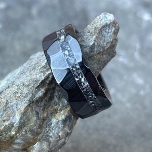 May include: A black tungsten carbide ring with a band of meteorite inlay. The ring has a hammered finish and is set on a gray rock.