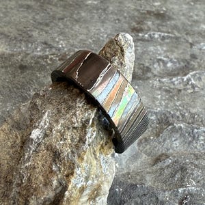 Pode incluir: Um anel largo de metal escuro com uma superfície texturizada e uma faixa de listras coloridas e iridescentes. O anel é exibido em uma pedra cinza áspera, mostrando seu design único. A largura do anel é de aproximadamente 1,3 cm.