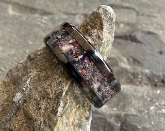 May include: A black ring with a rectangular shape and a colorful, speckled inlay. The inlay features a mix of purple, red, and white fragments. The ring is set against a textured, gray stone background.