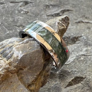 May include: A polished silver ring with a band featuring a mosaic of green and gold-toned segments. The ring is set against a textured gray rock background, highlighting its reflective surface and unique design.