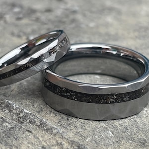 May include: Two matching silver rings with a dark, textured inlay. The rings have a faceted, hammered finish on the outer band. One ring is thinner than the other, suggesting a set for a couple.