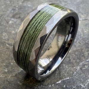 May include: A faceted, gunmetal-colored ring with a central band of tightly wound, olive-green material. The ring's design features a geometric pattern on the outer edges, creating a textured appearance. The ring is shown on a gray, textured surface.