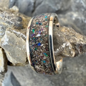 May include: A silver ring with a wide band featuring a speckled, multi-colored inlay. The inlay has a mix of brown, gray, blue, green, and purple hues. The ring is set against a natural, gray stone background.