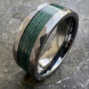 Op de afbeelding: Een zilverkleurige ring met een donkergroene middenband. De ring heeft een gefacetteerd ontwerp op de buitenranden, wat contrasteert met de gladde, gepolijste band. Het groene midden lijkt van geweven materiaal te zijn gemaakt.