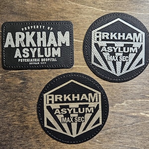 May include: Three black leather patches with white text and designs. The top patch reads "PROPERTY OF ARKHAM ASYLUM PSYCHIATRIC HOSPITAL GOTHAM CITY". The bottom two patches read "ARKHAM ASYLUM MAX SEC" with a stylized design.