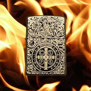 May include: A black and gold lighter with an ornate design and a Saint Benedict medal on the front. The lighter is in front of a background of flames.