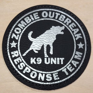 May include: Black leather patch with a silver outline and text. The text reads "Zombie Outbreak Response Team" with a star on either side. In the center is a silver silhouette of a dog with the text "K9 Unit" below.