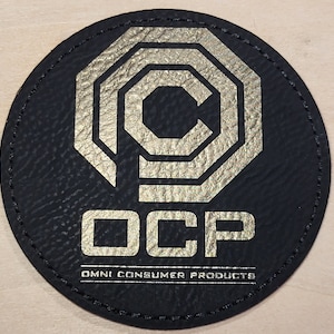 May include: A round, black patch with a gold-colored geometric design and the letters "OCP". The text "OMNI CONSUMER PRODUCTS" is printed below the letters. The patch has a stitched border.