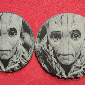 May include: Two round black slate coasters with a gray and white image of a character from a science fiction movie. The character has a large head with a wrinkled forehead, large eyes, and a serious expression.