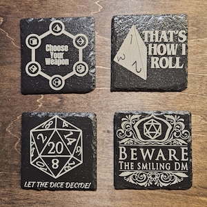 May include: Set of four black slate coasters with white designs. The coasters feature dice and game-related phrases, including "Choose Your Weapon", "That's How I Roll", "Let the Dice Decide!" and "Beware the Smiling DM".