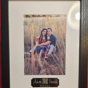 May include: Framed photograph featuring a couple embracing in a field of tall grass. The photo is set within a white mat and a black wooden frame. A small plaque at the bottom reads "Jan & Veronica" and "2-22-22".