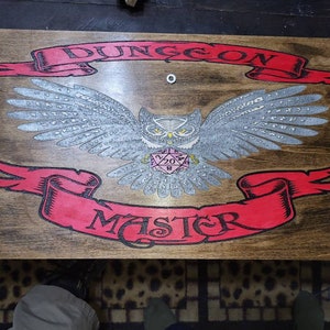 May include: Wooden game board with a brown finish, featuring a detailed owl illustration with outstretched wings. Red banners above and below read "DUNGEON" and "MASTER". A pink die is in the center. Decorative metal corner accents.