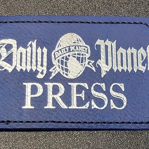 May include: A rectangular blue patch with the words "Daily Planet PRESS" in white, stylized lettering. The Daily Planet logo, a globe with lightning bolts, is centered above the word PRESS. The patch has a black stitched border.