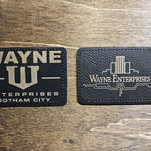 May include: Two black leather coasters with gold lettering. The first coaster says "WAYNE ENTERPRISES GOTHAM CITY" with a stylized "W" logo. The second coaster says "WAYNE ENTERPRISES" with a stylized cityscape graphic.