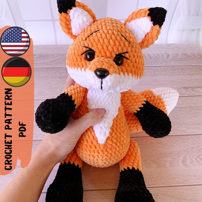 Op de afbeelding: Gehaakte oranje en witte pluche vos met zwarte accenten. Het speelgoed heeft een witte buik en zwarte ogen. Het speelgoed zit op een houten oppervlak.